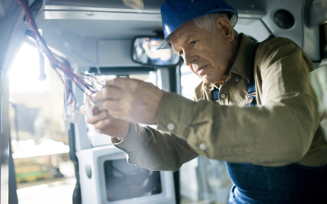 Concentrated senior male industrial electrician in hardhat connecting naked electric wires while working inside of machine cab